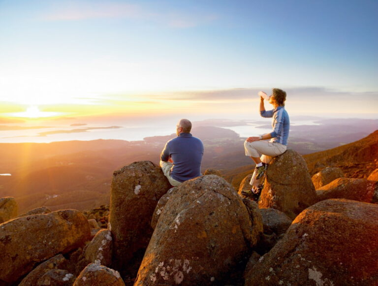 A couple sit on the boulders of Mt Wellington, looking at the sun set over the city of Hobart