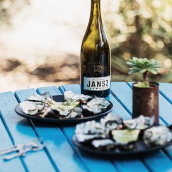 A painted wooden table with 2 plates of shucked oysters and a bottle of Jansz bubbly.