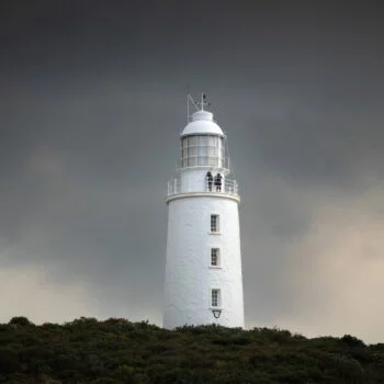Cape Bruny Lighthouse. A stark white stone lighthouse with 2 people on the observation deck. Bushes in the foreground and ominous cloudy sky background.